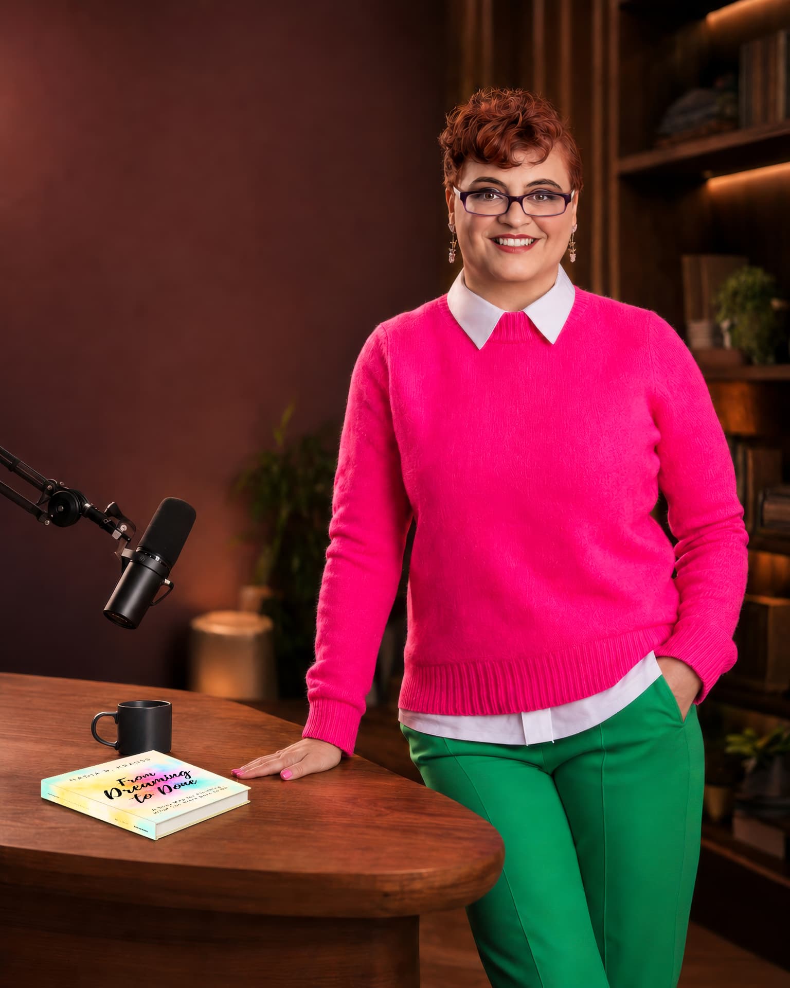 Nadia in a hot-pink sweater and green trousers beside her book From Dreaming to Done and a microphone